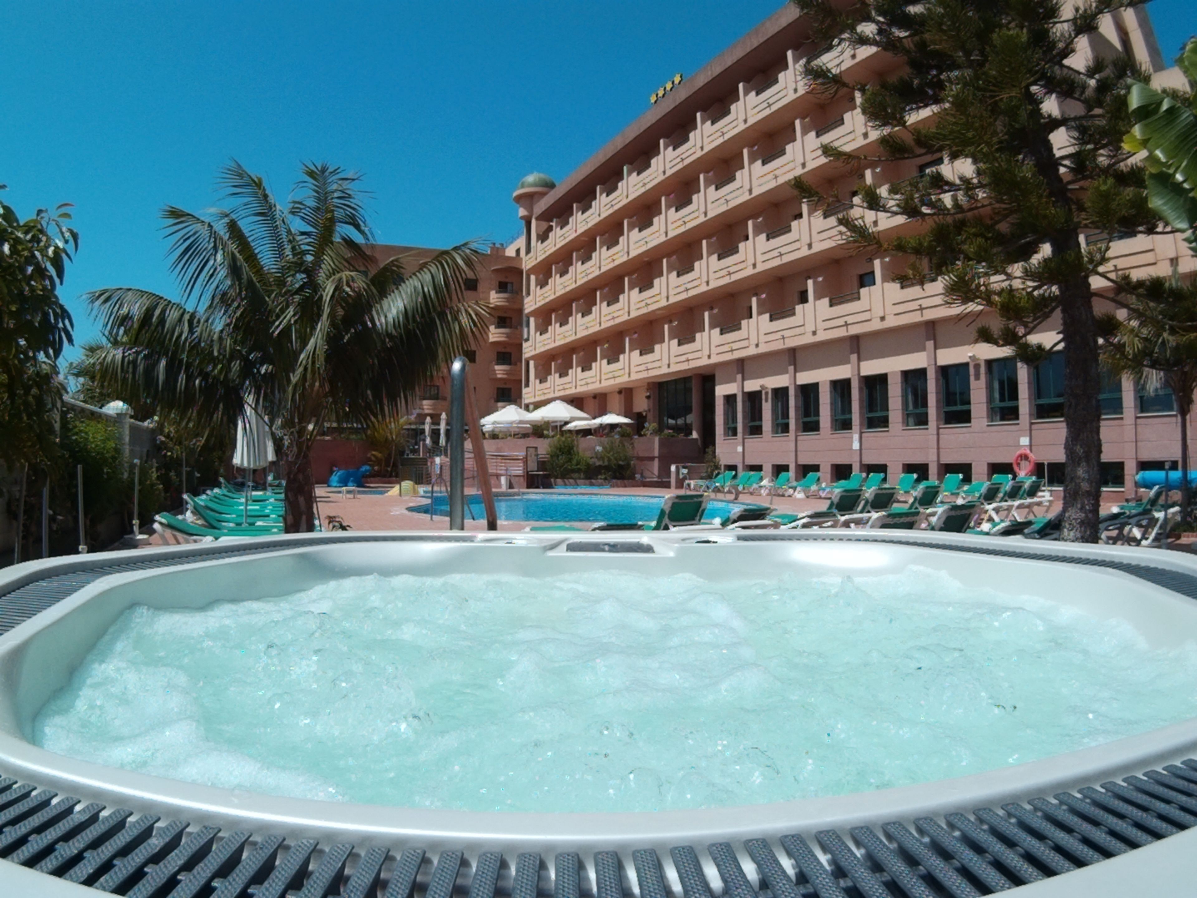A hot tub area with a pool and lounge chairs, surrounded by palm trees and a multi-story building with balconies.