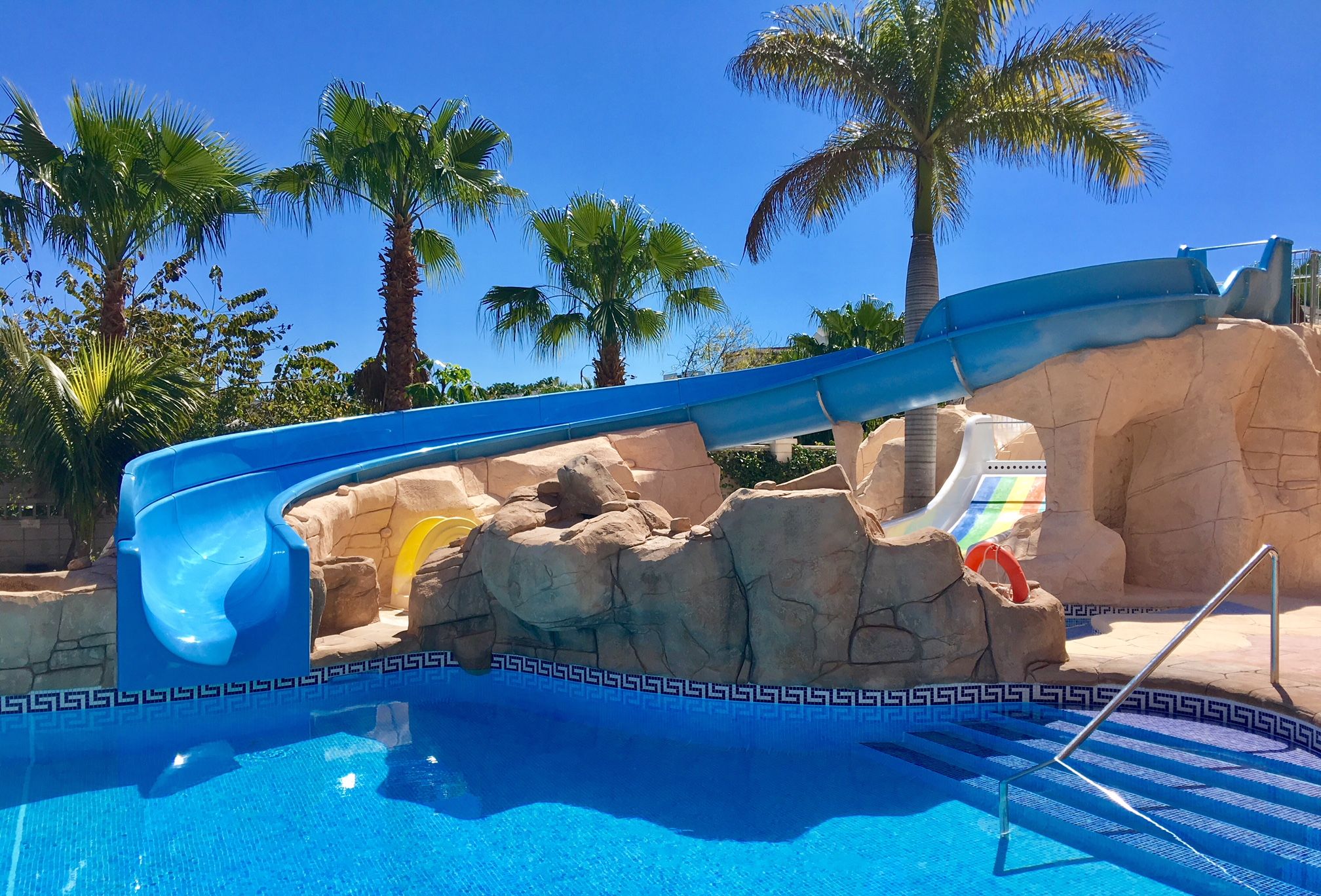 A pool with a slide, palm trees, and a clear blue sky.