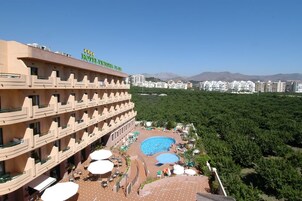 Un hotel con varios balcones, piscina y vistas a un paisaje urbano con montañas al fondo.
