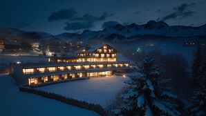 A multi-story hotel with a prominent central building and several smaller structures, surrounded by snow-covered mountains and a lake. - La Casies Mountain Living Hotel (Valle di Casies)