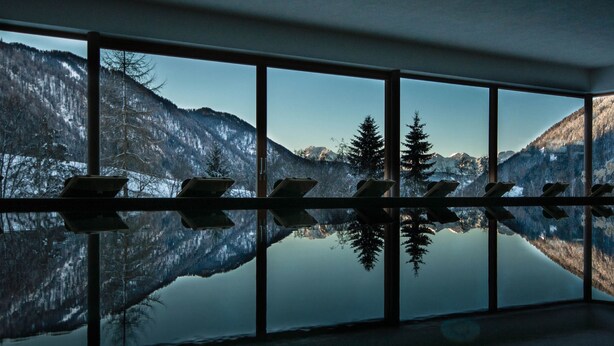 A room with a large window reflecting a mountainous landscape and a calm lake. - La Casies Mountain Living Hotel (Valle di Casies)