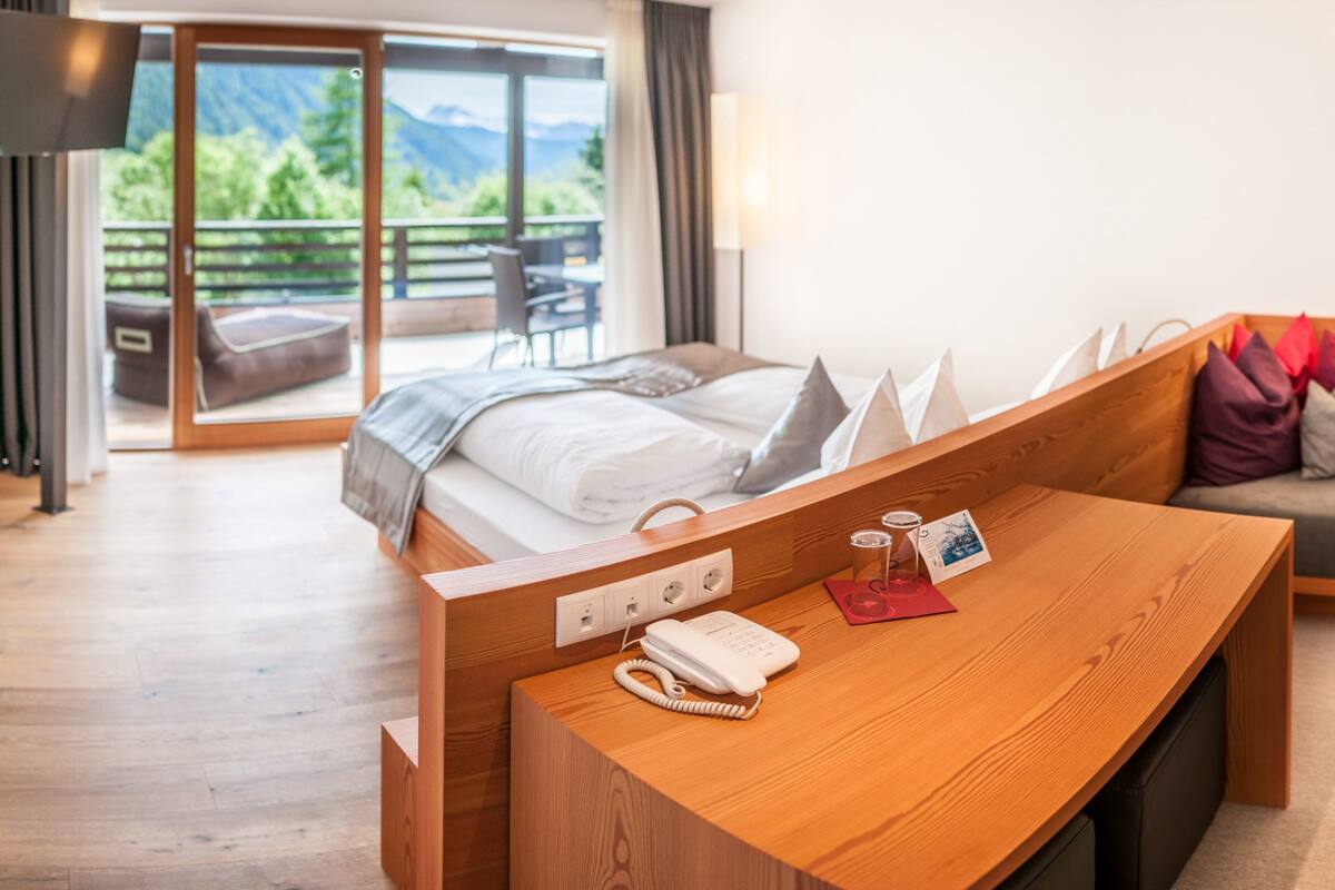 A hotel room with a bed, a wooden desk with a phone and glasses, a sofa with cushions, and a view of mountains through a sliding door.