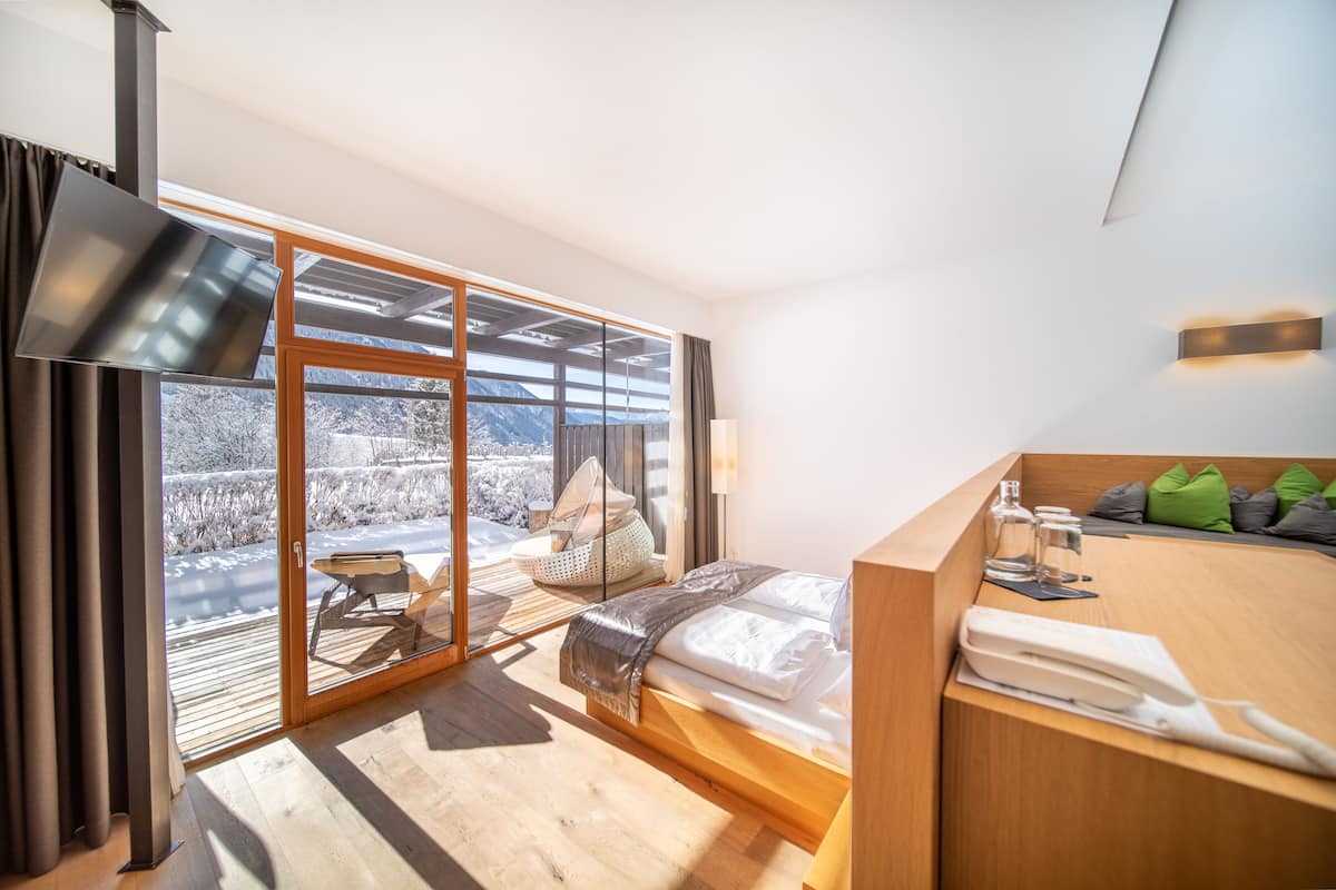 A modern hotel room with a large sliding glass door leading to a balcony, a wooden bed, a wooden desk, and a view of a snowy landscape.