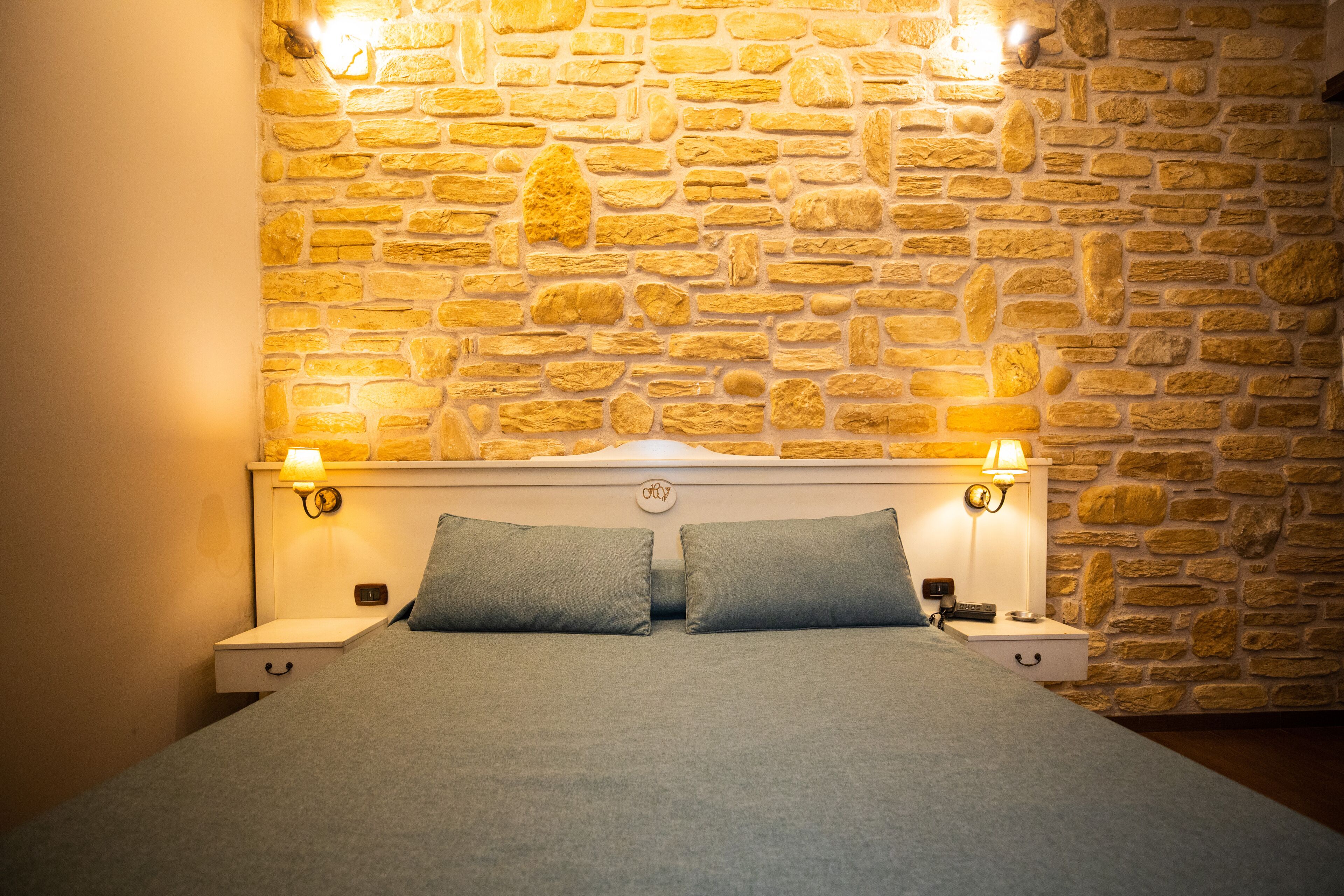 A bed with a stone wall backdrop, two bedside lamps, and a nightstand with a lamp.