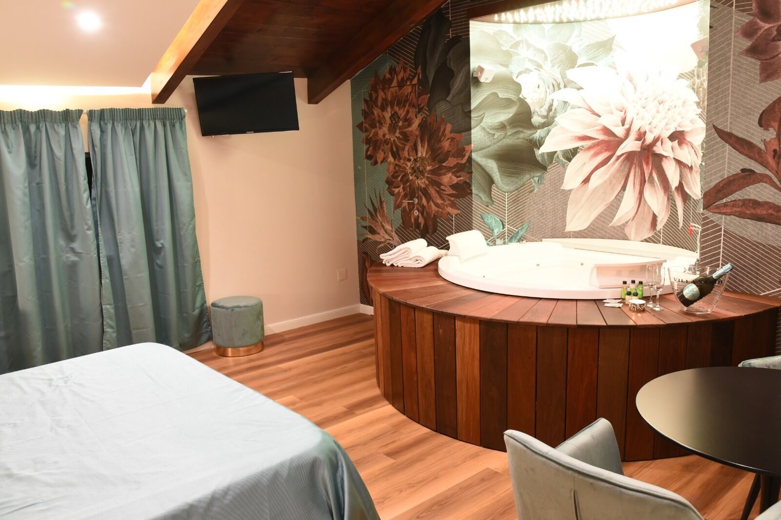 A bedroom with a bed, a round wooden table, a small stool, and a wall-mounted TV.