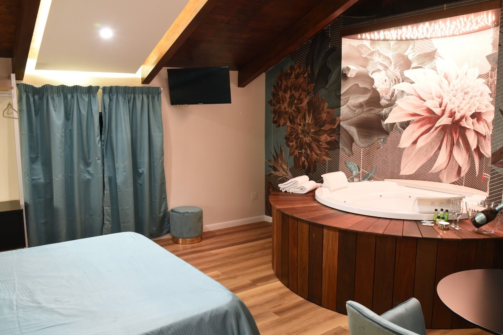 Habitación de hotel con cama, tocador de madera redondo con bañera, un taburete pequeño y televisor en la pared.