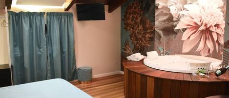 Habitación de hotel con cama, tocador de madera redondo con bañera, un taburete pequeño y televisor en la pared.
