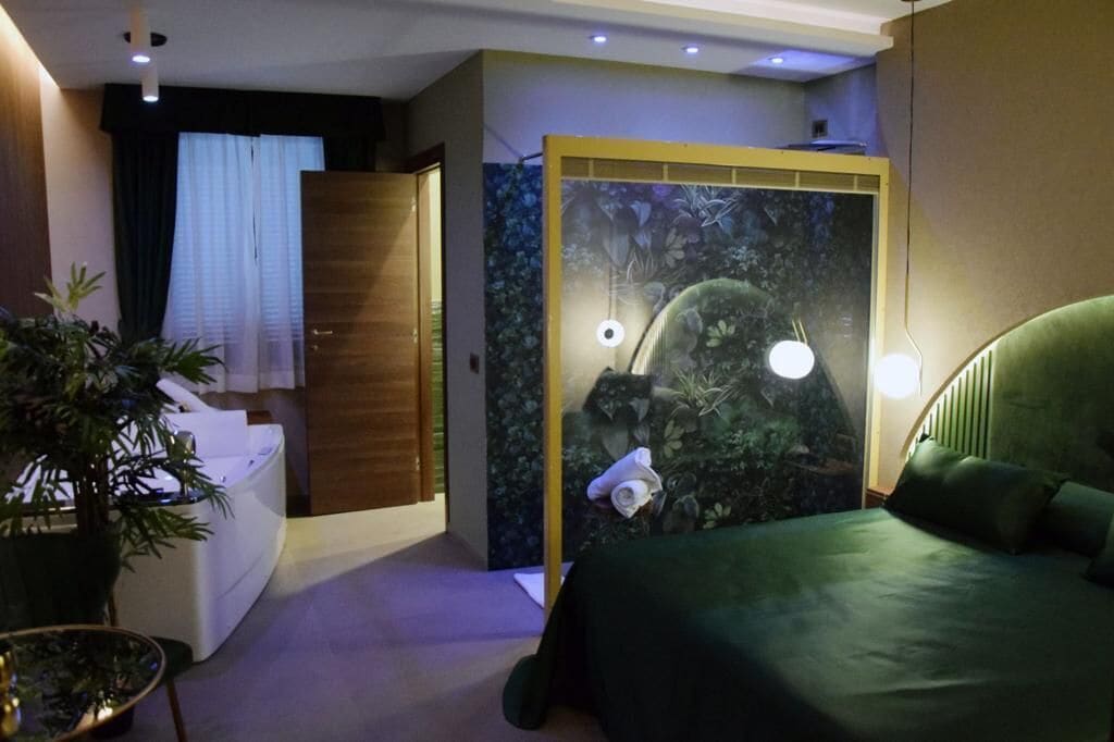 A hotel room with a bed, a small desk, a plant, and a wall with a nature-themed mural.