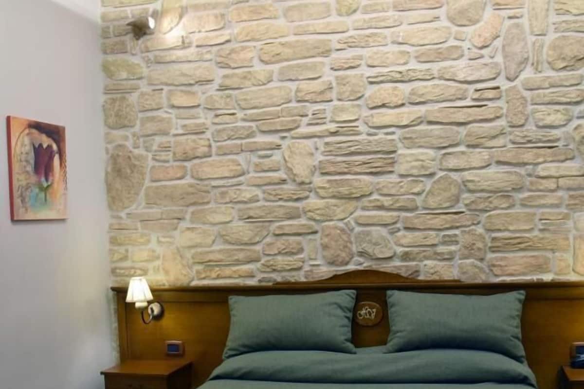 A bedroom with a stone wall, a bed with green bedding, a nightstand with a lamp, and a small table.