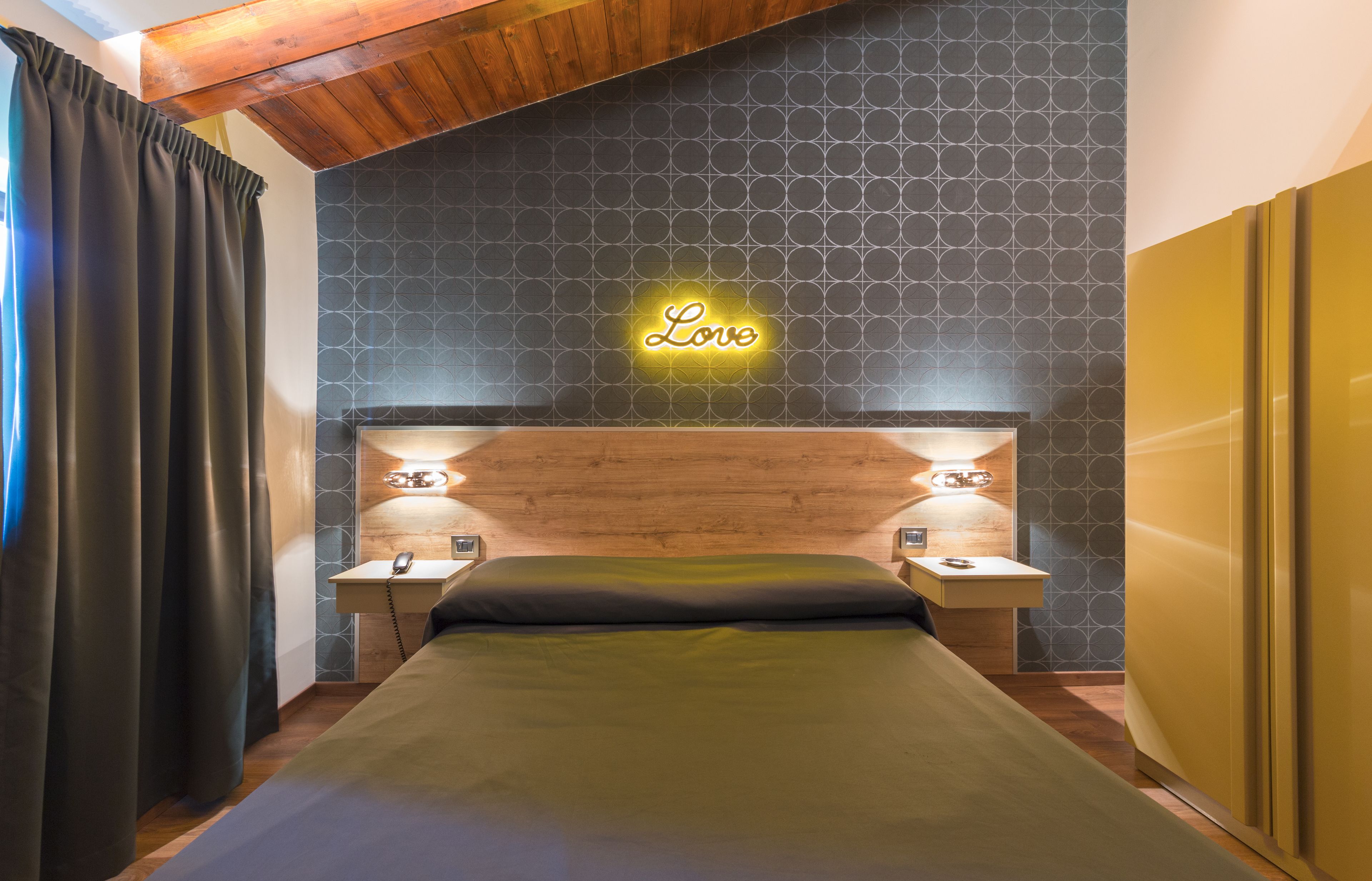 A hotel room with a bed, a neon 'Love' sign on the wall, wooden headboard, bedside tables with lamps, and a curtain on the left.
