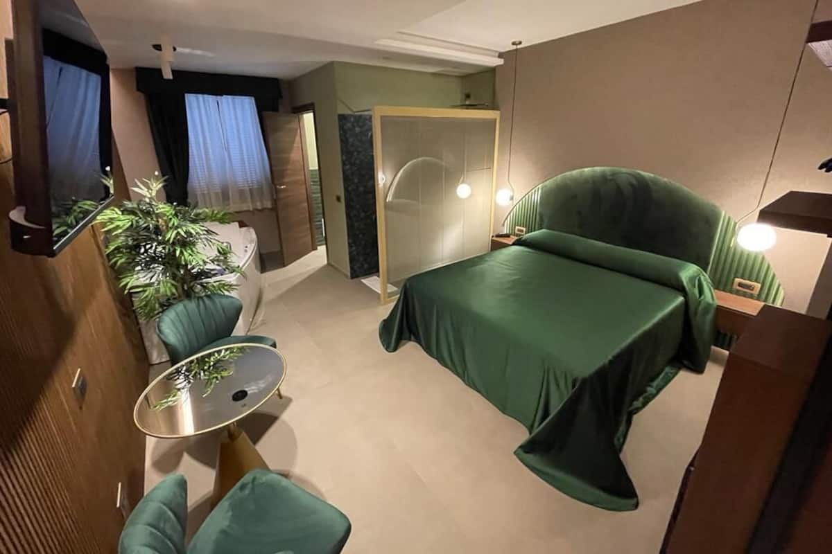 A modern hotel room with a green bed, a small round table, a green armchair, and a potted plant.