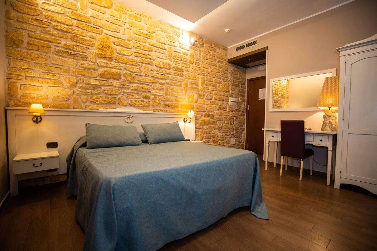 A bedroom with a stone wall, a bed with a blue bedspread, a nightstand, a desk, and a wardrobe.