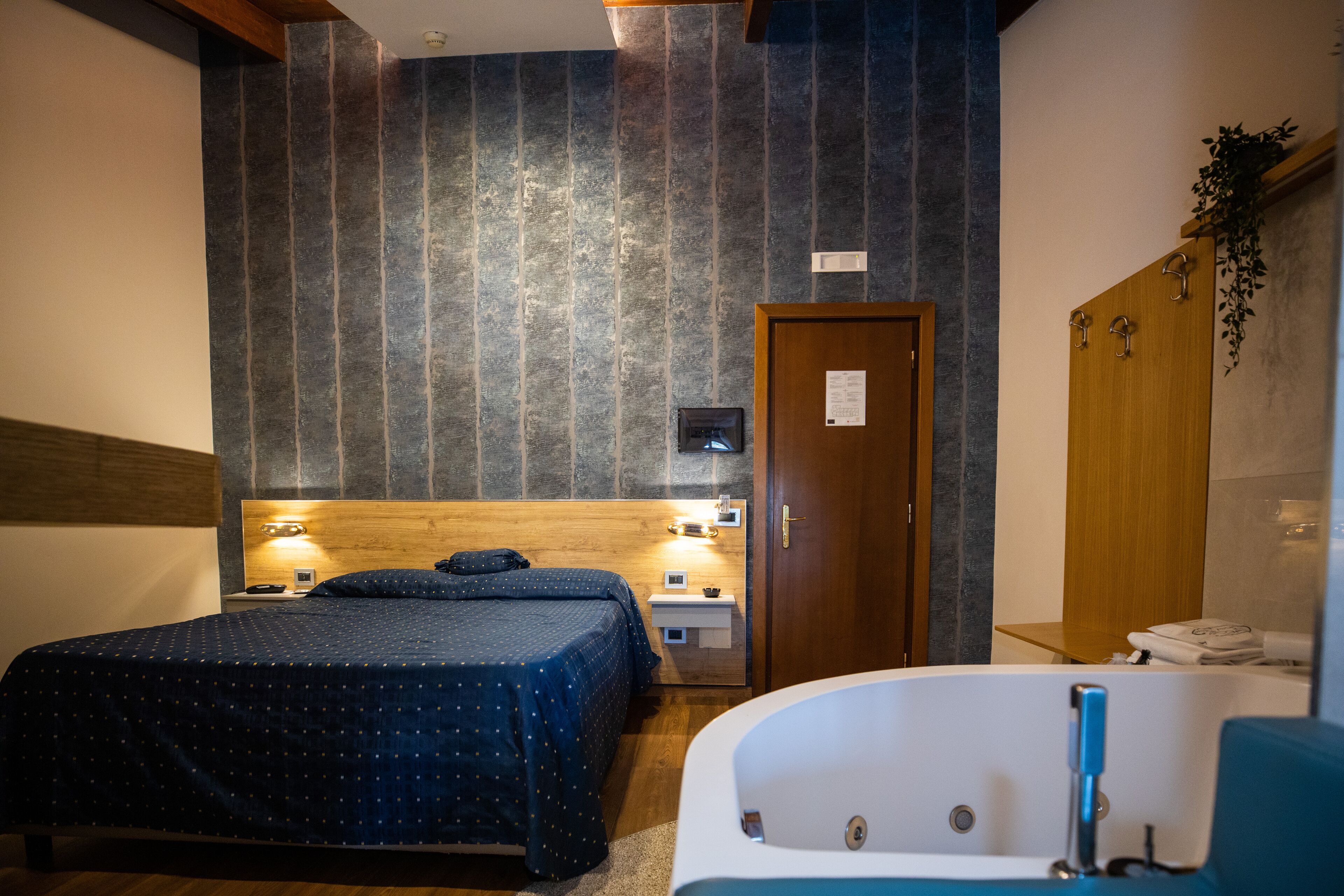 Un dormitorio con cama, cabecera de madera, mesita de noche, televisor montado en la pared, puerta y baño con bañera y perchero para toallas.
