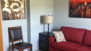 Una sala de estar con un sofá rojo, una silla de madera, una mesita auxiliar negra con una lámpara y dos cuadros en las paredes.