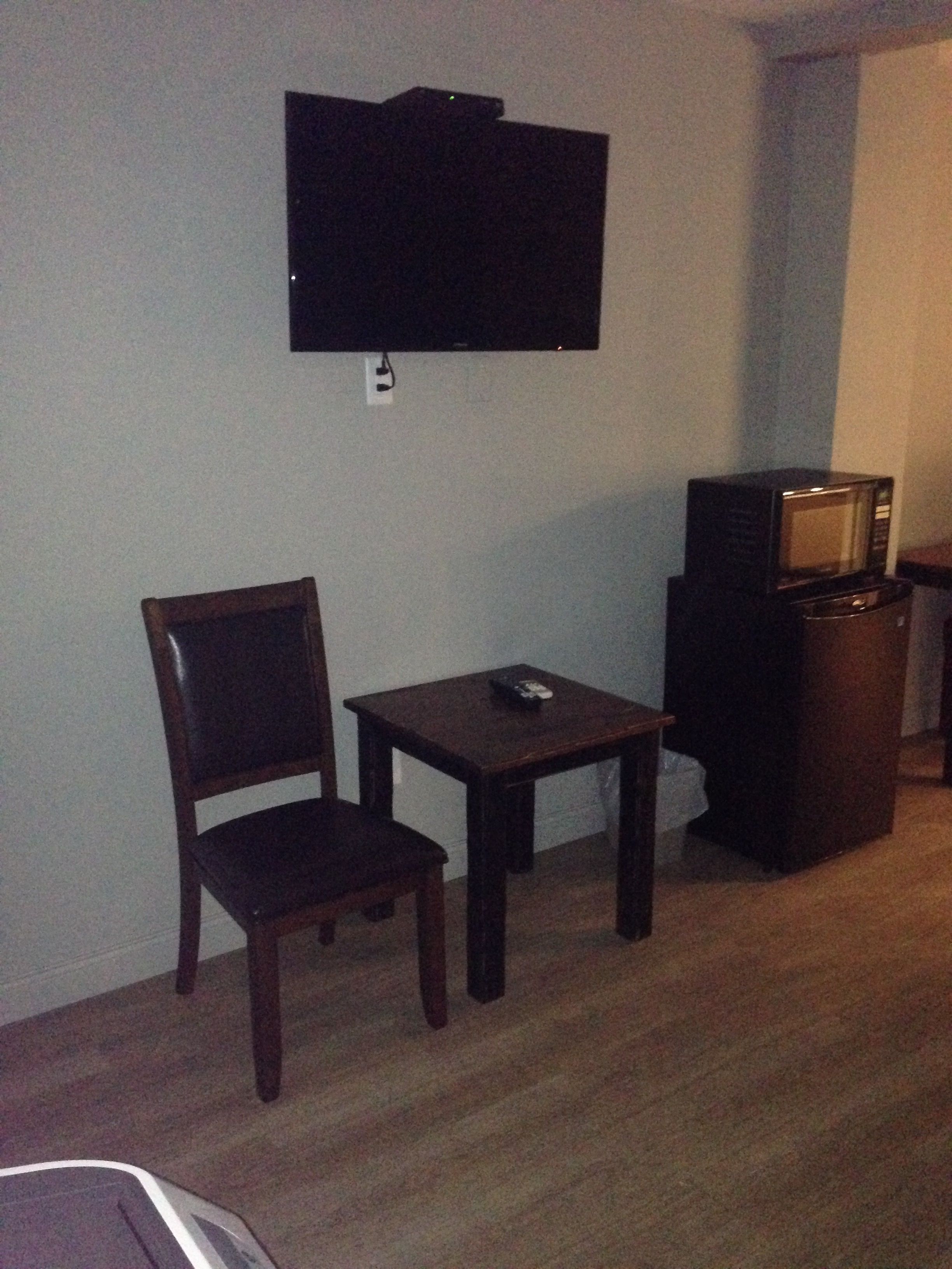 A small room with a television mounted on the wall, a wooden chair, a small table with a remote control, and a microwave on a cabinet.