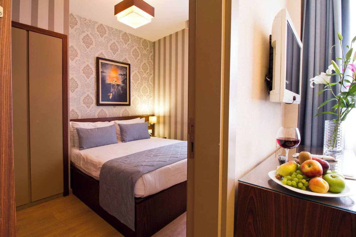 Ein Hotelzimmer mit einem Bett, einem Nachttisch mit Obstteller, einem Fernseher und einem gerahmten Bild an der Wand.