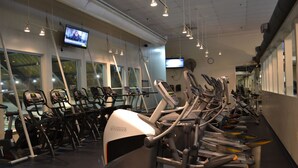 Un gimnasio con diferentes máquinas de ejercicio, una pantalla de televisión y amplias ventanas.