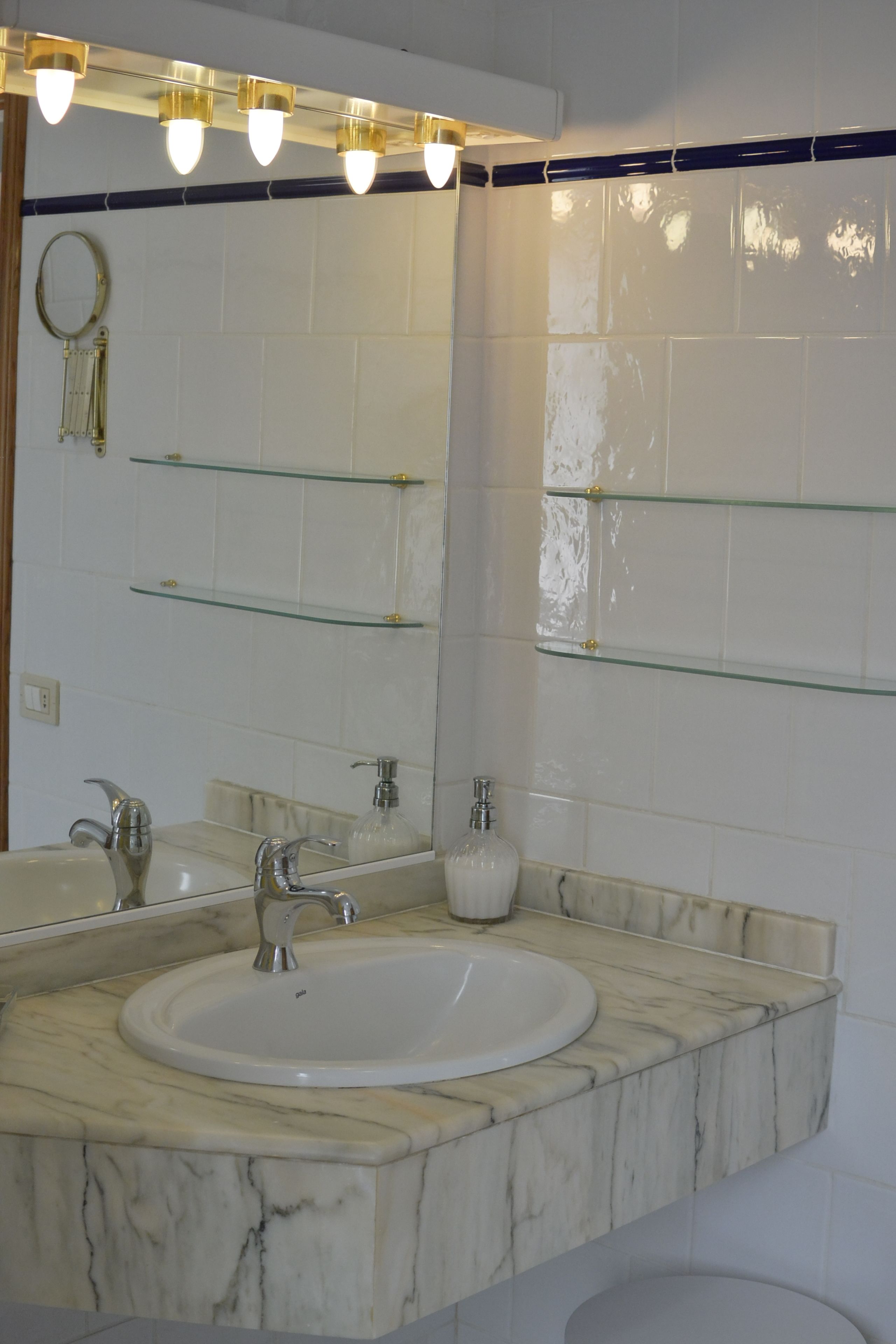 Un baño con lavamanos de mármol, repisas de vidrio y un espejo.