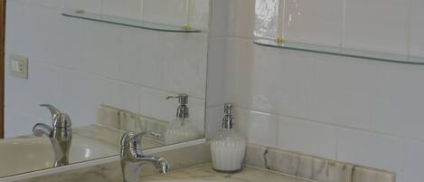 Un baño con lavamanos de mármol, repisas de vidrio y un espejo.