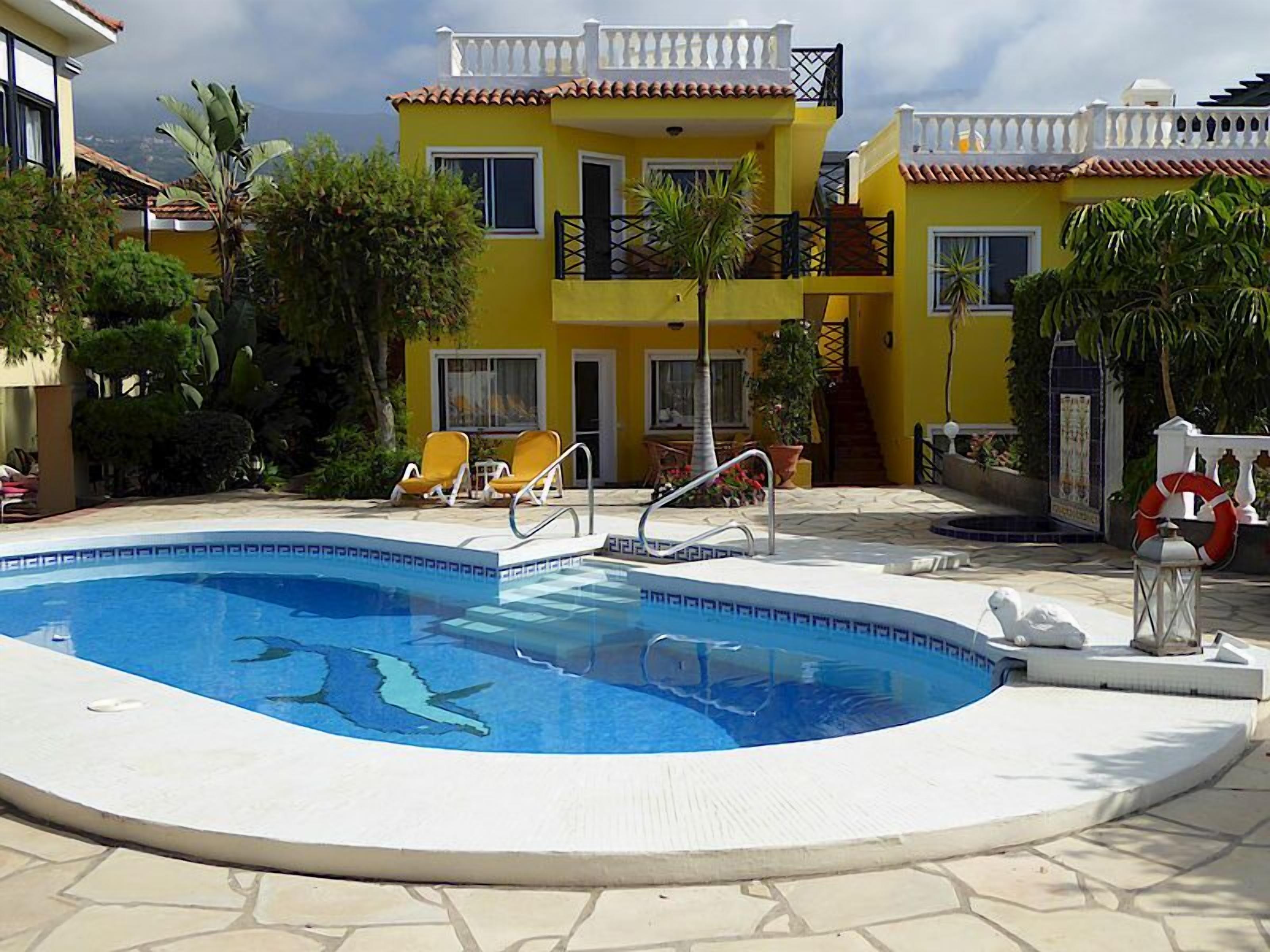 A swimming pool with two lounge chairs, a lifebuoy, and a staircase leading to a yellow building with a balcony.