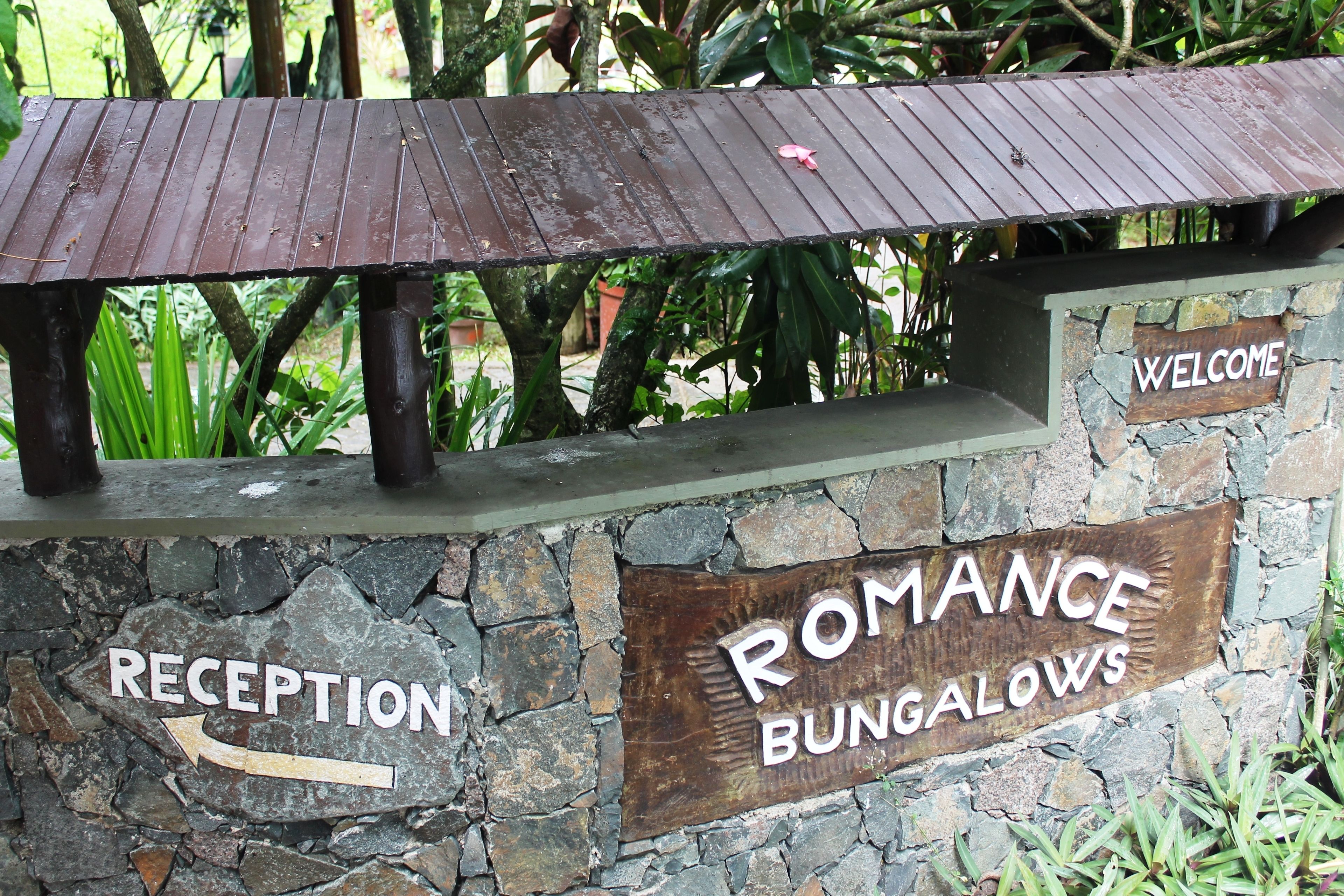 Un espace d’accueil avec un banc en bois et un mur en pierre. On y trouve des panneaux indiquant « RÉCEPTION » et « BUNGALOWS ROMANTIQUES ».