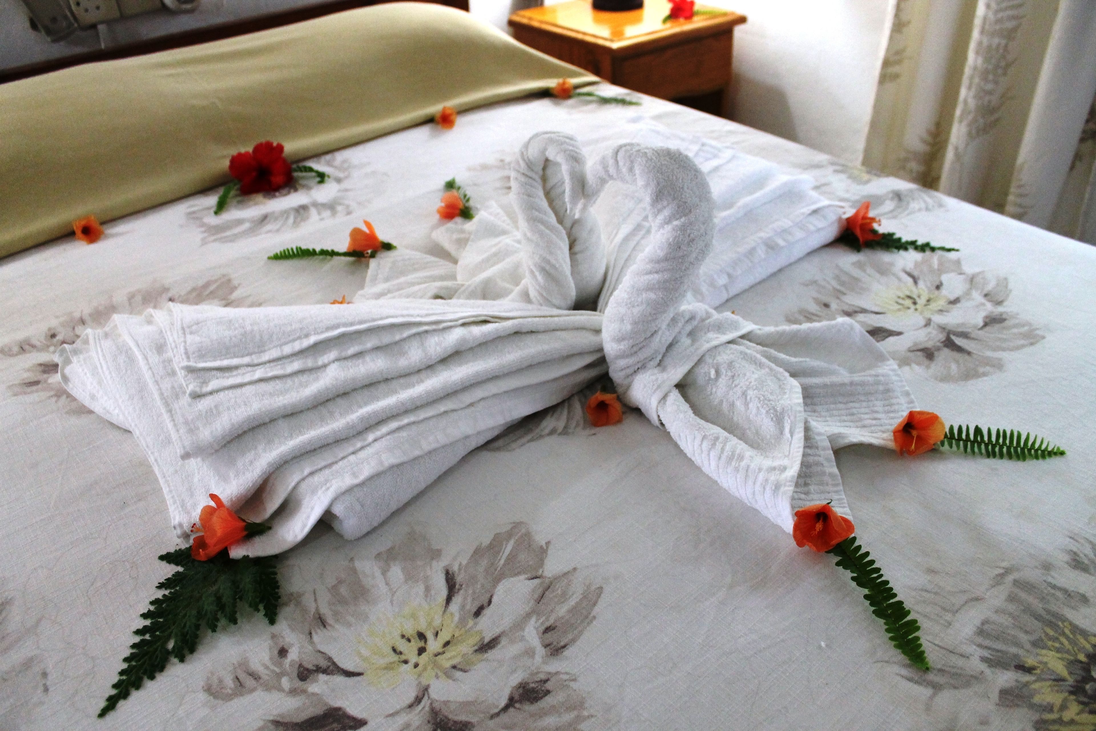 Ein ordentlich bezogenes Bett mit floralem Bettbezug und Handtüchern, die als Schwan angeordnet sind, verziert mit orangefarbenen Blumen und grünen Blättern.