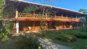Um edifício de madeira de dois andares com varanda, rodeado por vegetação e um céu claro.