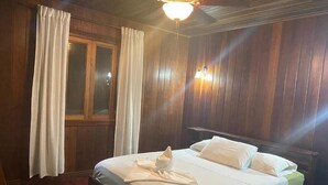 Uma cama bem arrumada com roupa de cama e toalha brancos, paredes de madeira, uma janela com cortinas e um lustre.