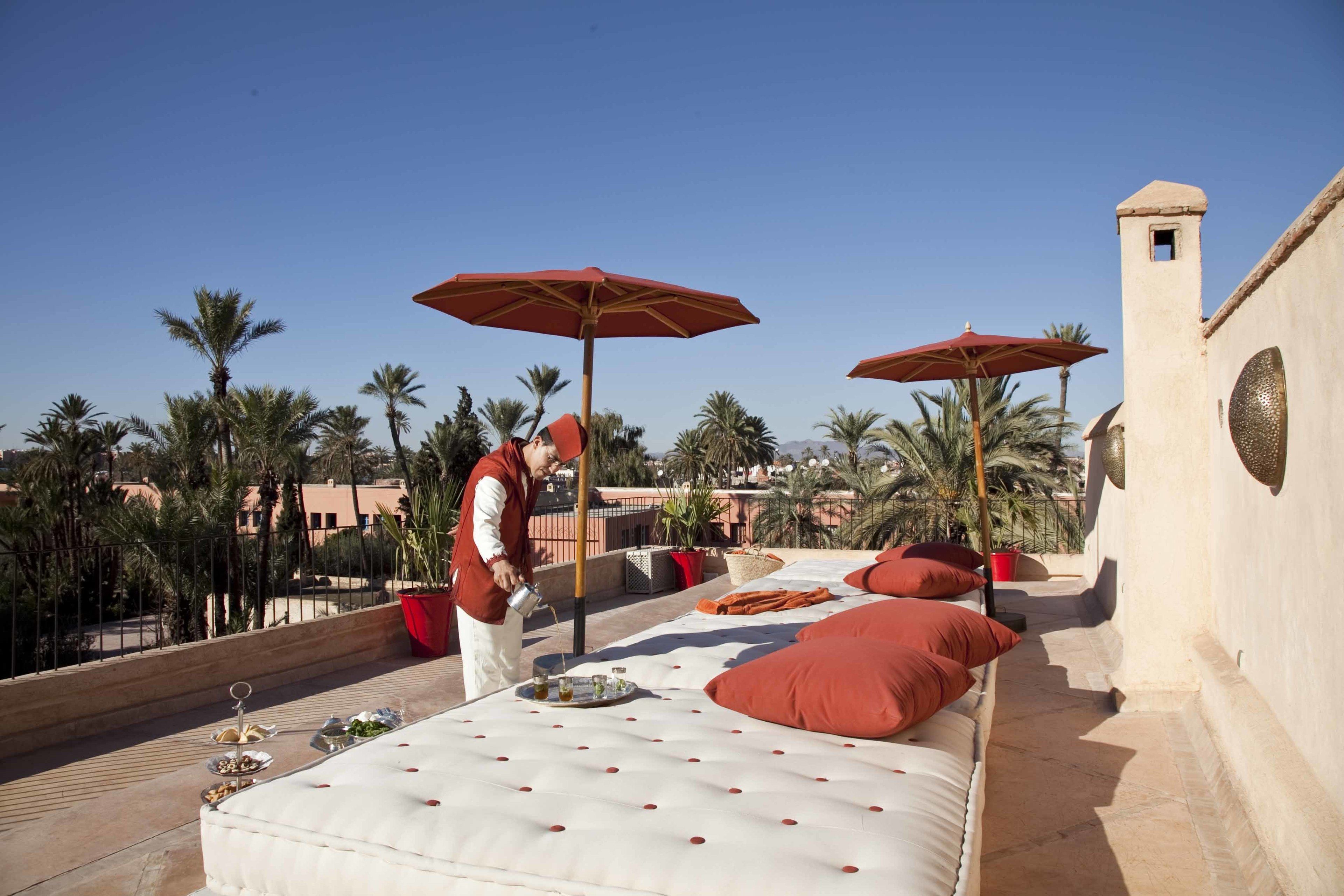 A rooftop terrace with a bed, red umbrellas, and palm trees.