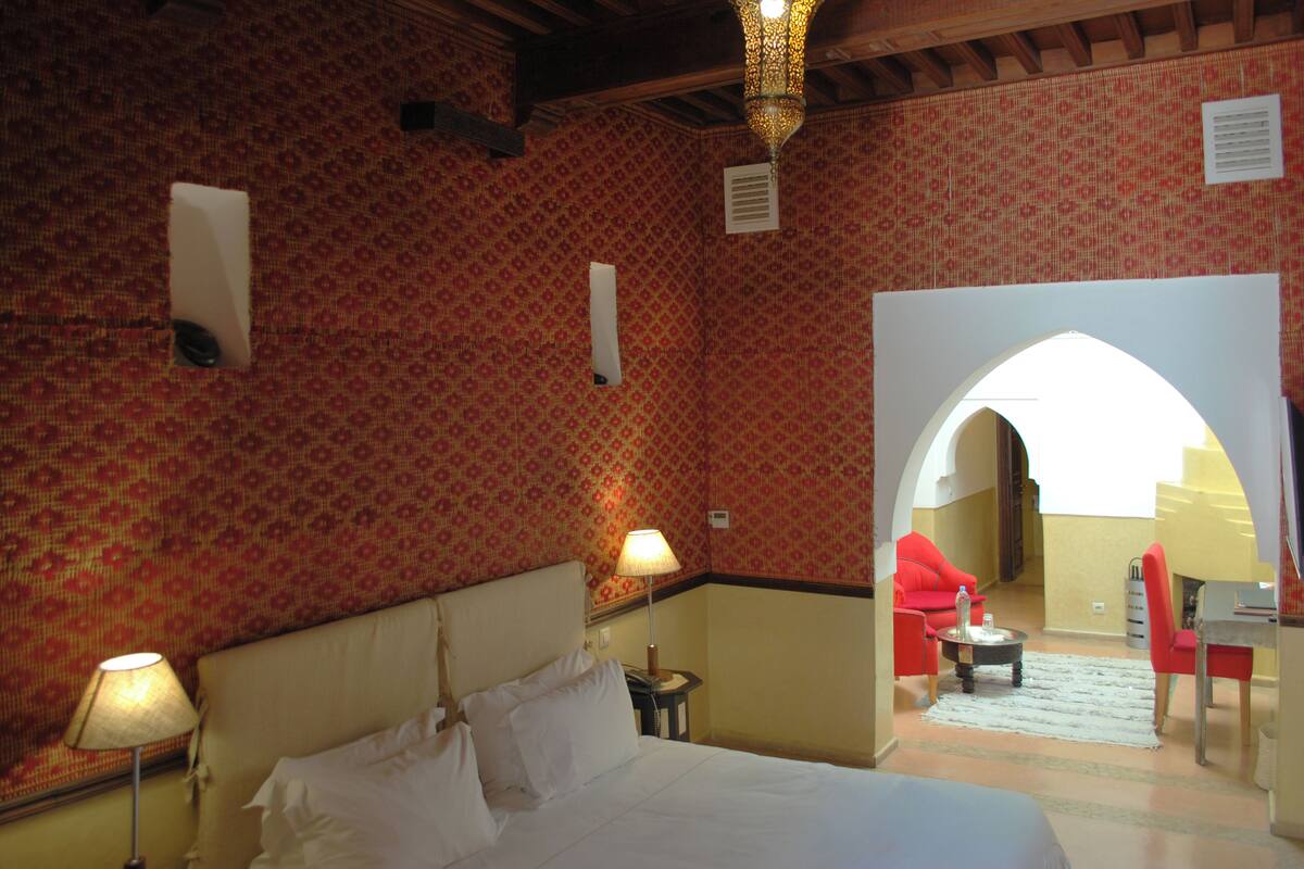 Habitación de hotel con cama, dos lámparas, escritorio y silla. Pared con diseño y un arco que conduce a otra habitación.