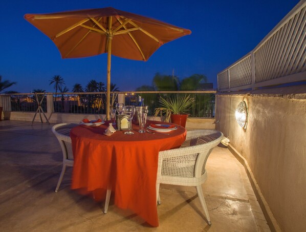 Un área de comedor al aire libre con una mesa preparada para dos, una silla de mimbre, una planta en maceta y un paraguas grande.