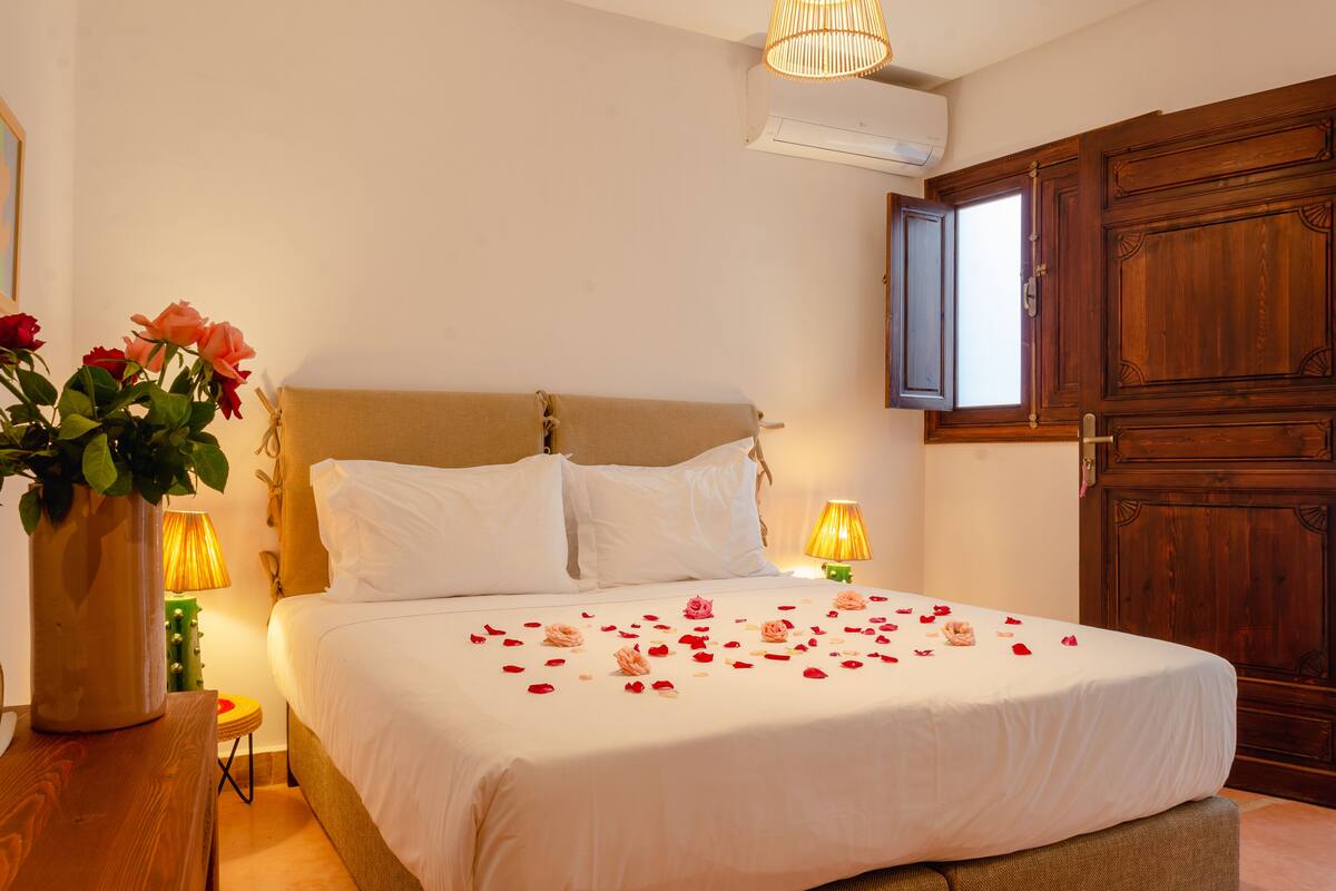 Una cama bien hecha con sábanas blancas y pétalos de rosa, una mesita de noche de madera con un florero y un armario de madera.