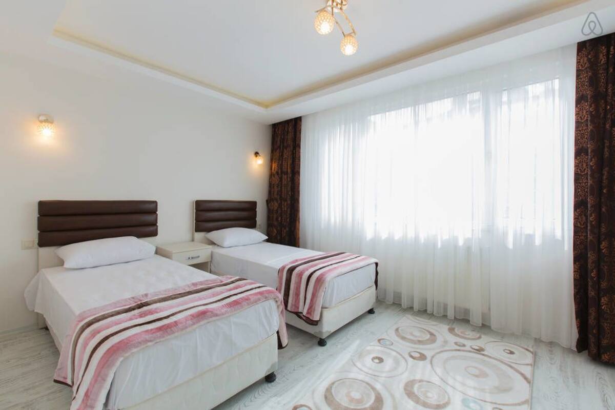 A hotel room with two beds, a large window with sheer curtains, a patterned rug, and a ceiling light fixture.