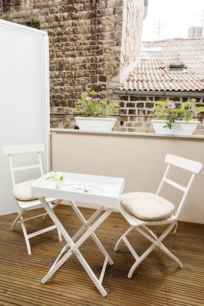 Zona exterior amb seients, taula blanca i dues cadires blanques, envoltada per plantes en testos i una paret de pedra.