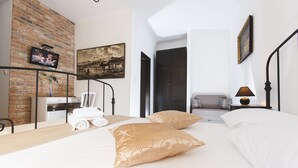 A modern hotel room with a brick wall, a flat-screen TV, a painting, a bed with white and gold pillows, a desk with a lamp, and a view of a cityscape through a window. - Prior Suites Split (Split)