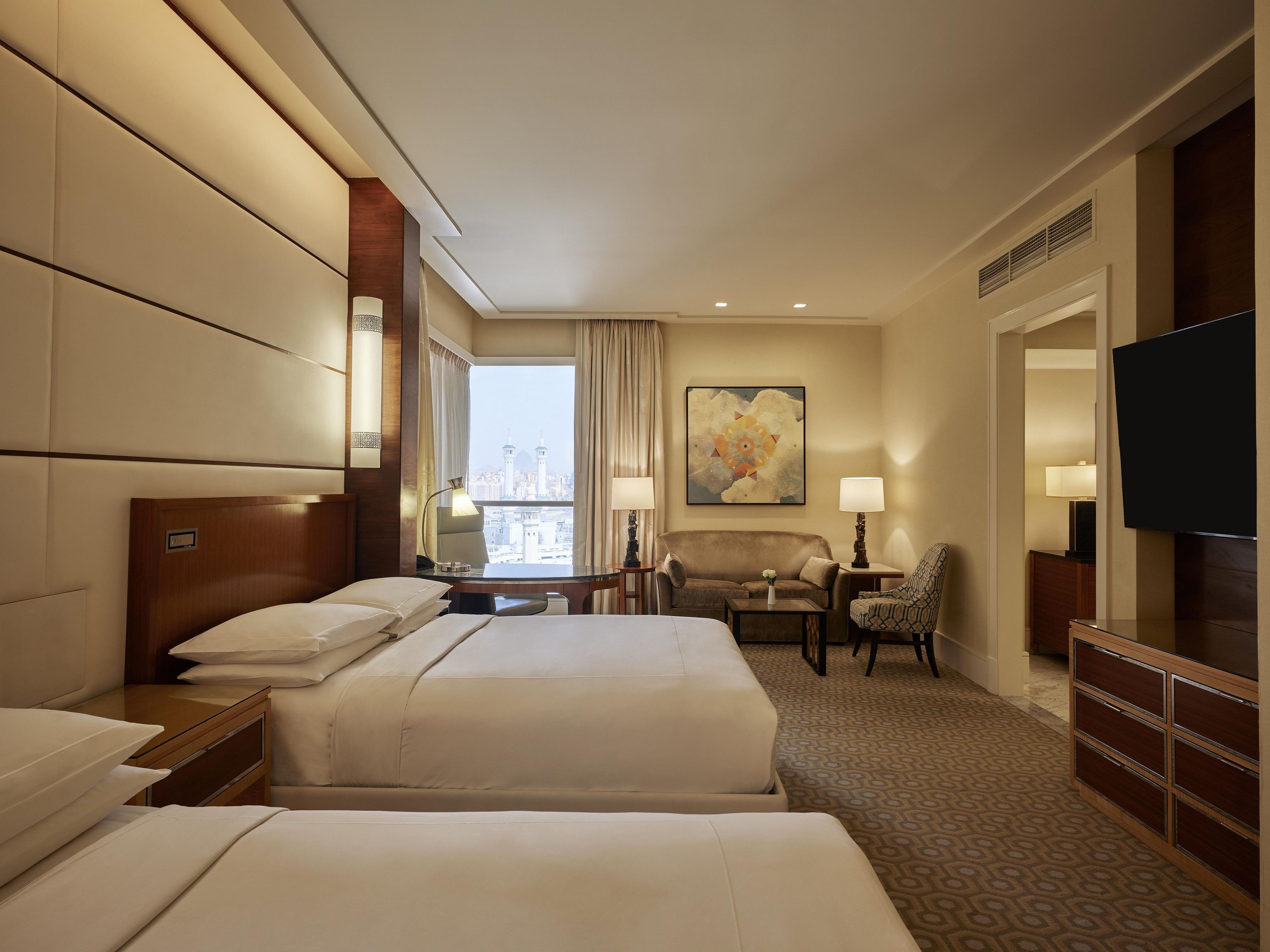 A hotel room with two beds, a desk, a chair, a TV, and a large window with a city view.
