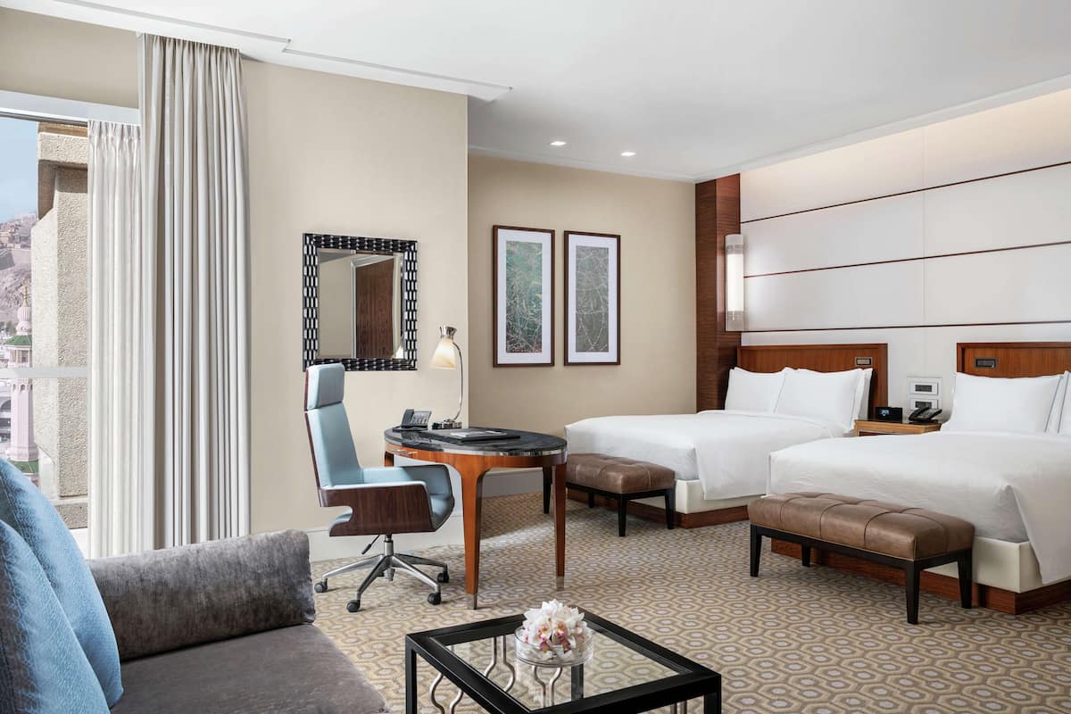 A hotel room with two beds, a desk, a chair, a coffee table, and a large window with curtains.
