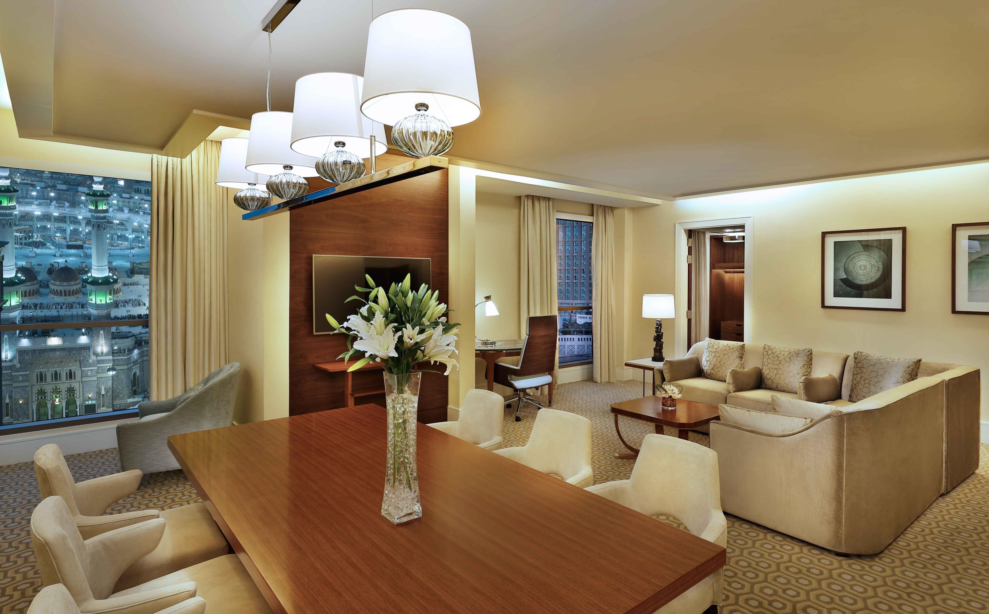 A hotel room with a dining table, sofa, chairs, a TV, and a view of a cityscape.