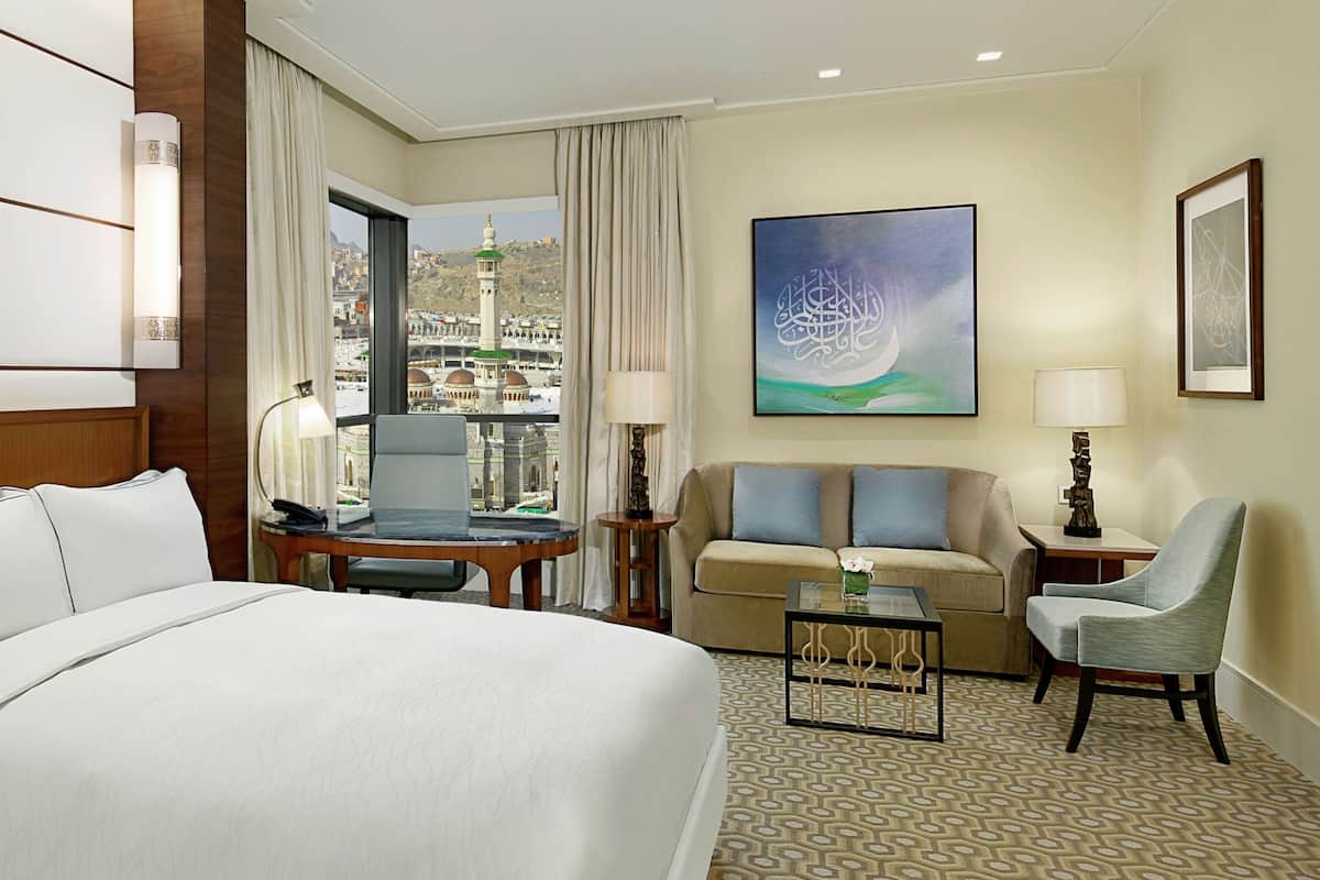 A hotel room with a large bed, a sofa, a chair, a small table, and a view of a cityscape with a mosque.