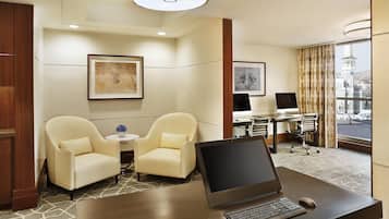 A hotel room with a desk, two armchairs, a small table with a vase, and a large window with a view of a tower.