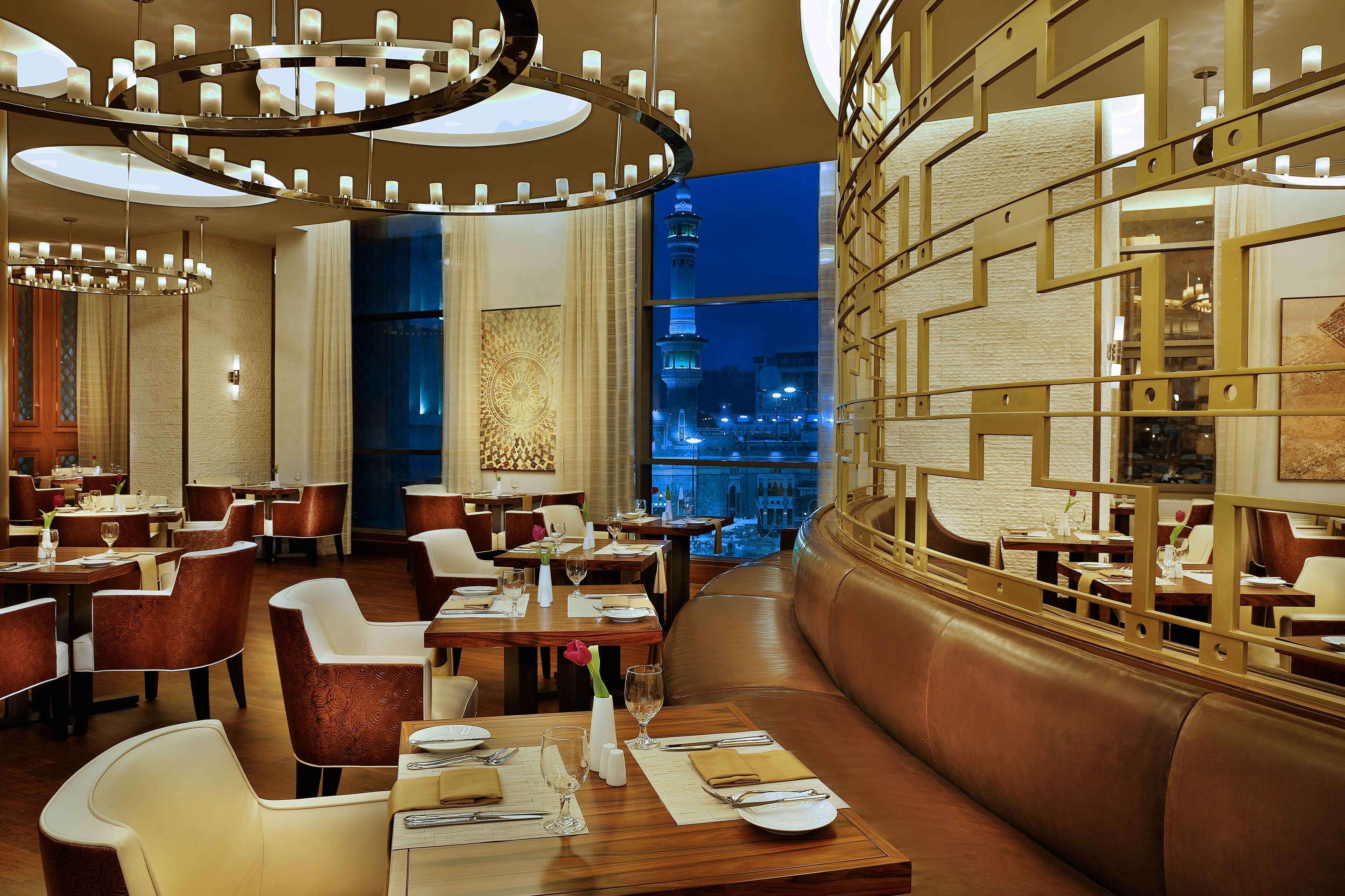 A modern restaurant with multiple dining tables and booths, a view of a cityscape at night, and a decorative wall with geometric patterns.