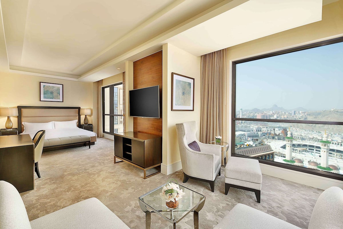 A hotel room with a bed, a TV, a desk, a chair, a sofa, and a view of a cityscape.