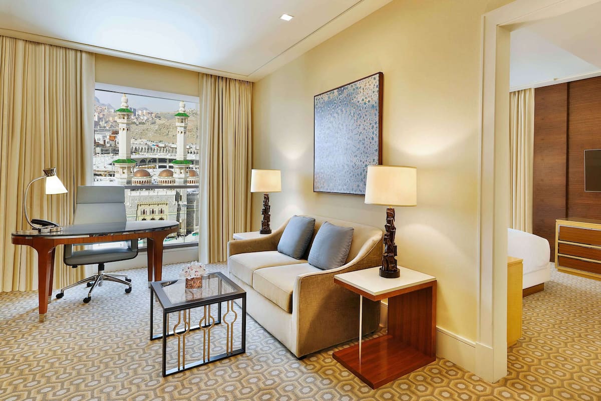 A hotel room with a sofa, a small table, a desk, a chair, a TV, and a view of a cityscape with a mosque.