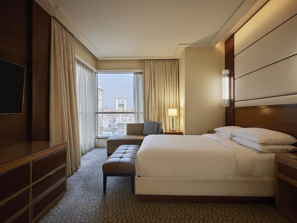 A hotel room with a large bed, a seating area with a chair and ottoman, a TV on the left, and a view of a cityscape through the window.