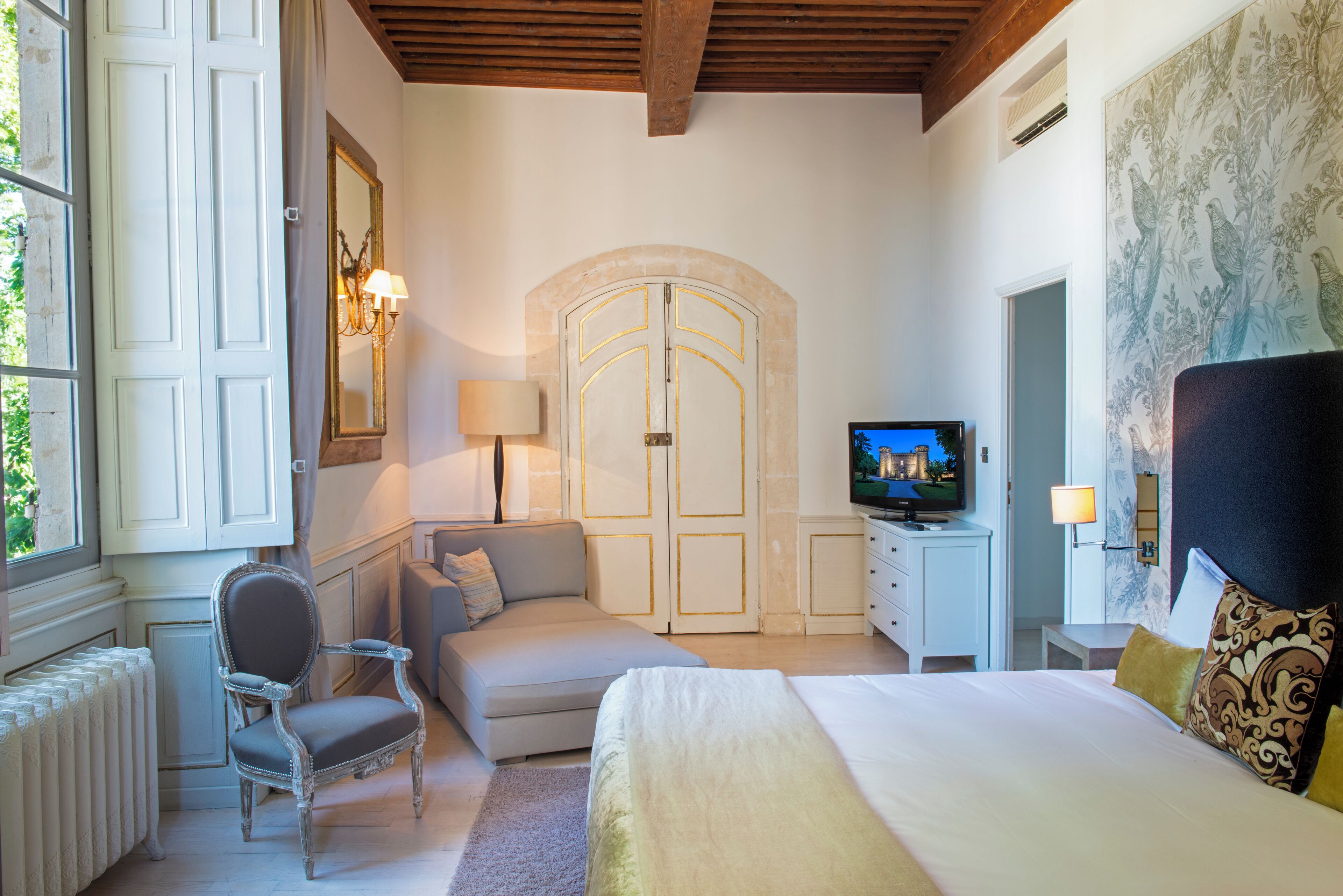 A bedroom with a bed, a sofa, a chair, a TV, and a large arched door.