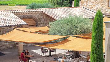 Un espace extérieur aménagé en terrasse, avec des murs en pierre, des tables et des chaises en bois, et des auvents orange qui offrent de l’ombre.