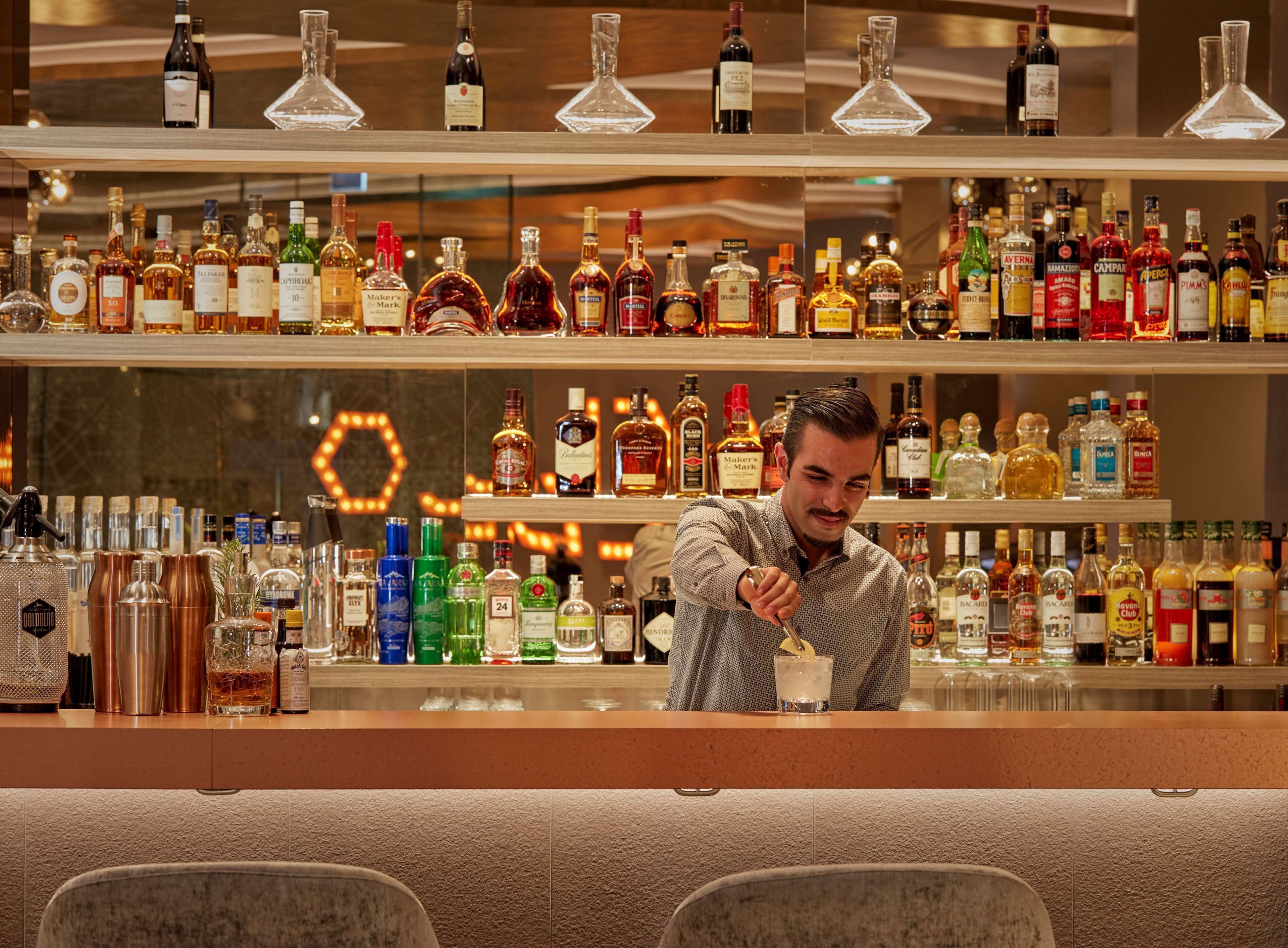 Un bar con varias estanterías llenas de botellas de licores, un barman preparando una bebida y una lámpara con forma hexagonal al fondo.