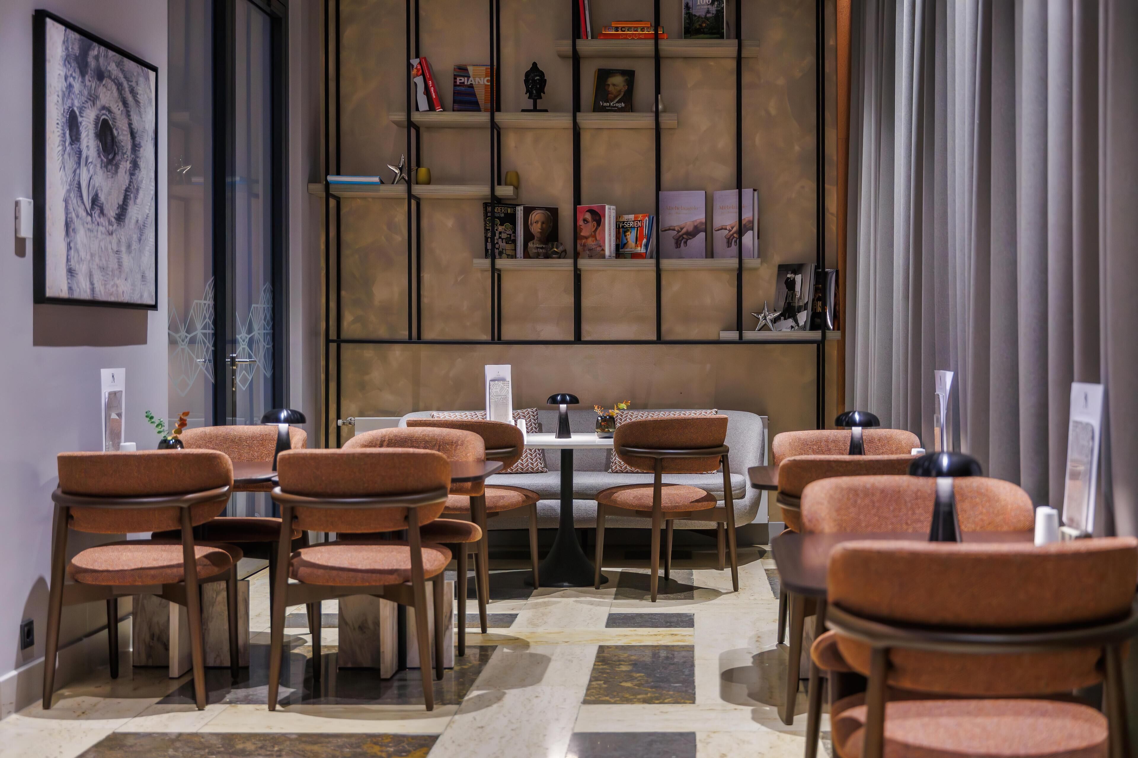 A modern hotel lobby with a central seating area, a bookshelf with books and decorative items, and a large window with curtains.