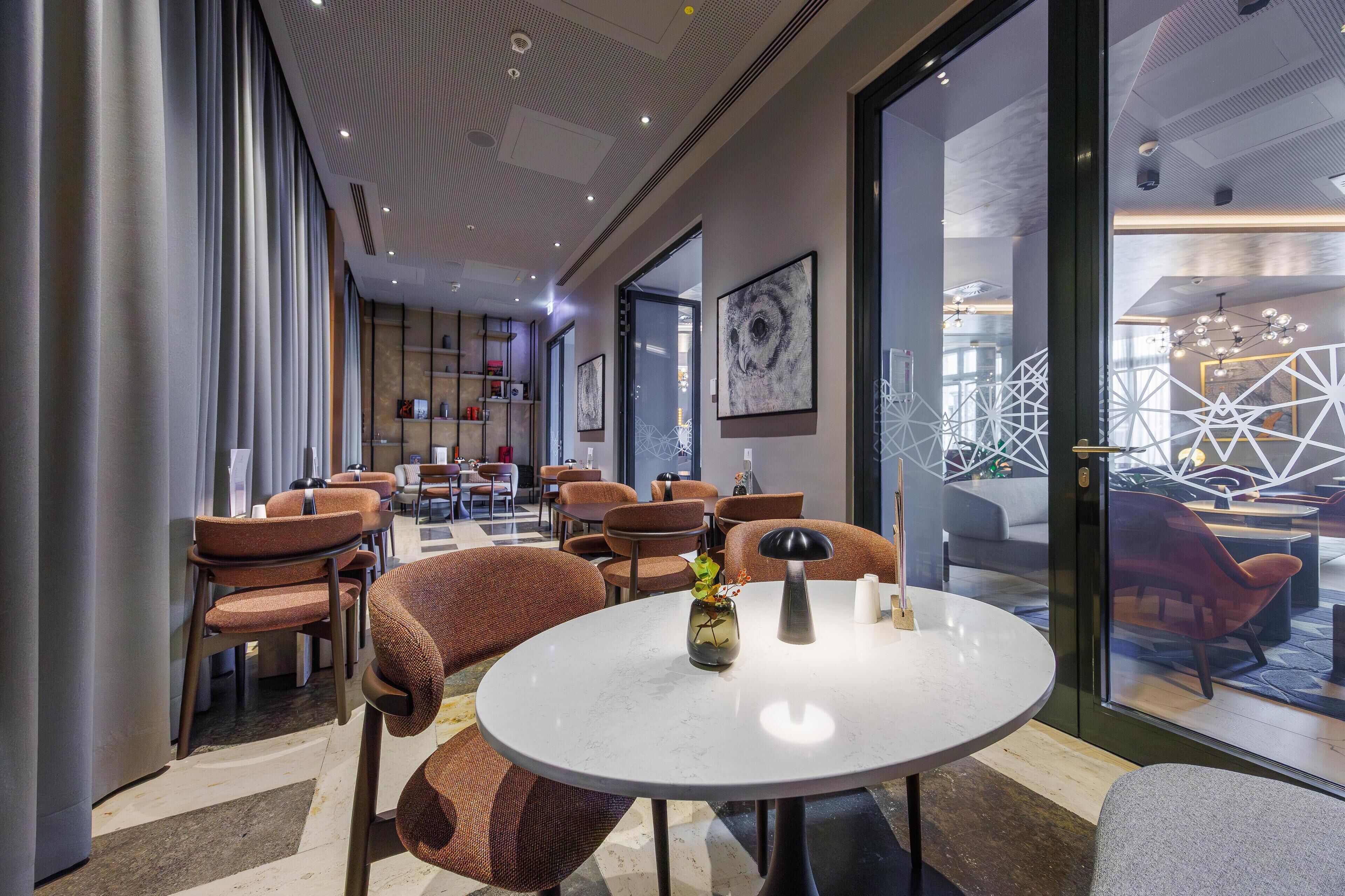 A modern interior with round tables and orange chairs, a glass wall with geometric decor, and a ceiling with recessed lighting.