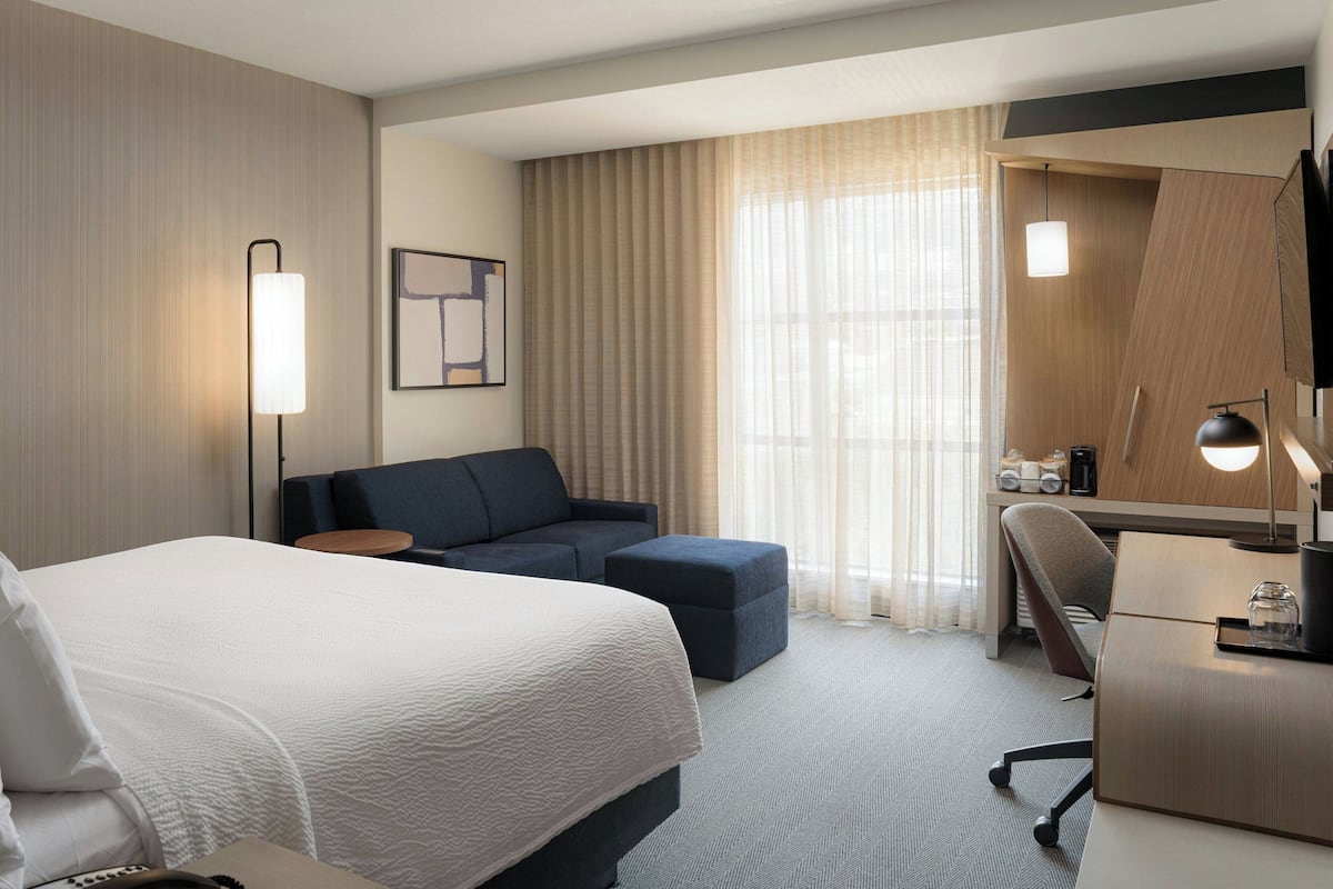 Ein Hotelzimmer mit einem großen Bett, einer Couch, einem Schreibtisch und einem Sessel. Ein Fenster mit Vorhängen, eine Lampe und ein gerahmtes Bild sind an der Wand angebracht.